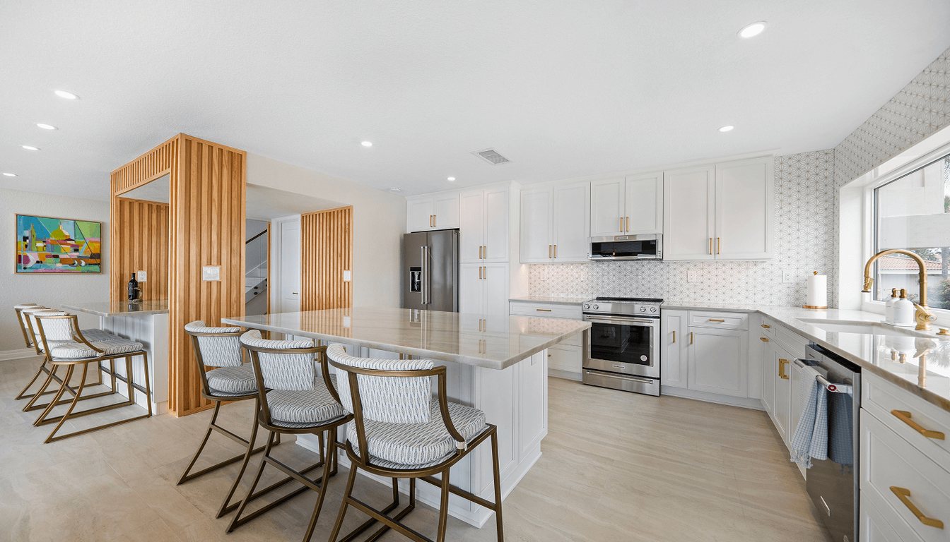Remodeled kitchen with white shaker cabinets, brass pulls, a stone-topped island with counter stools, patterned tile backsplash and slatted wood partitions