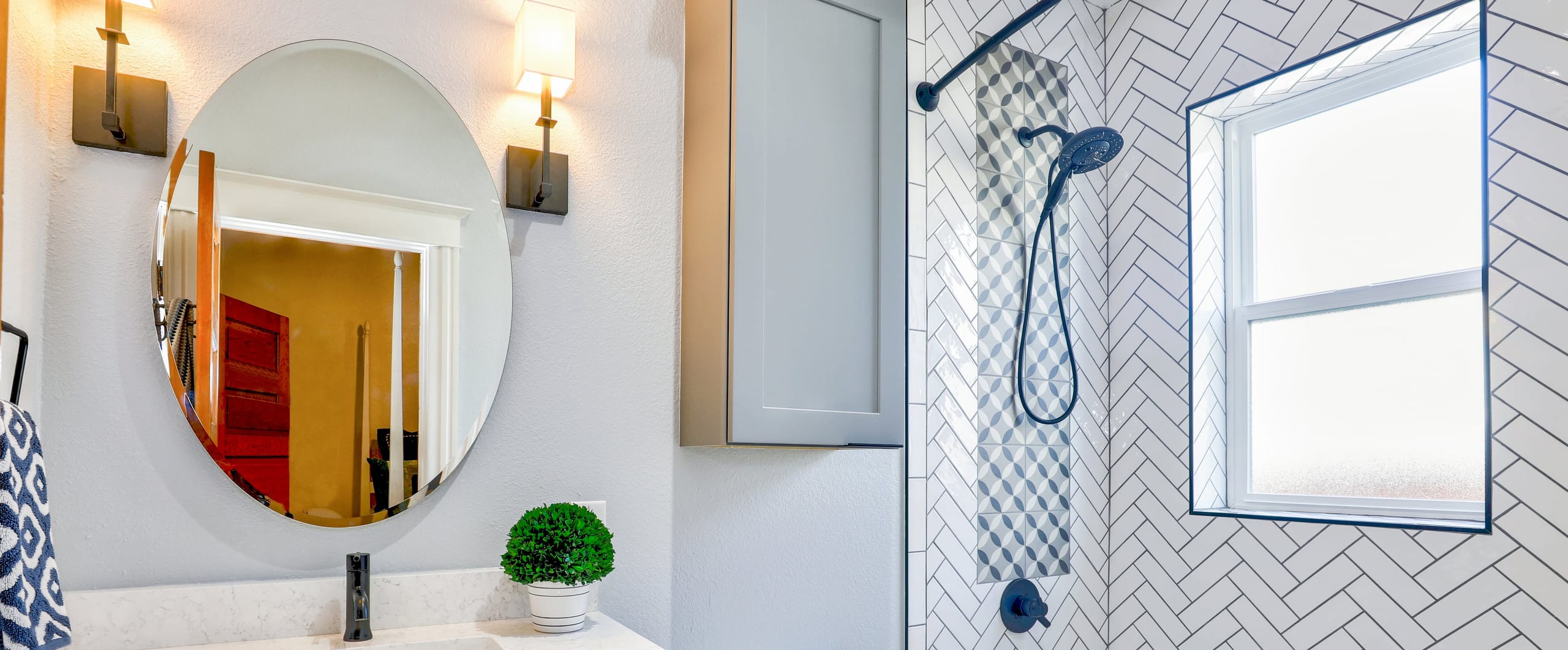 Bright bathroom with a round mirror between brass sconces, a quartz vanity, and a shower tiled in white herringbone