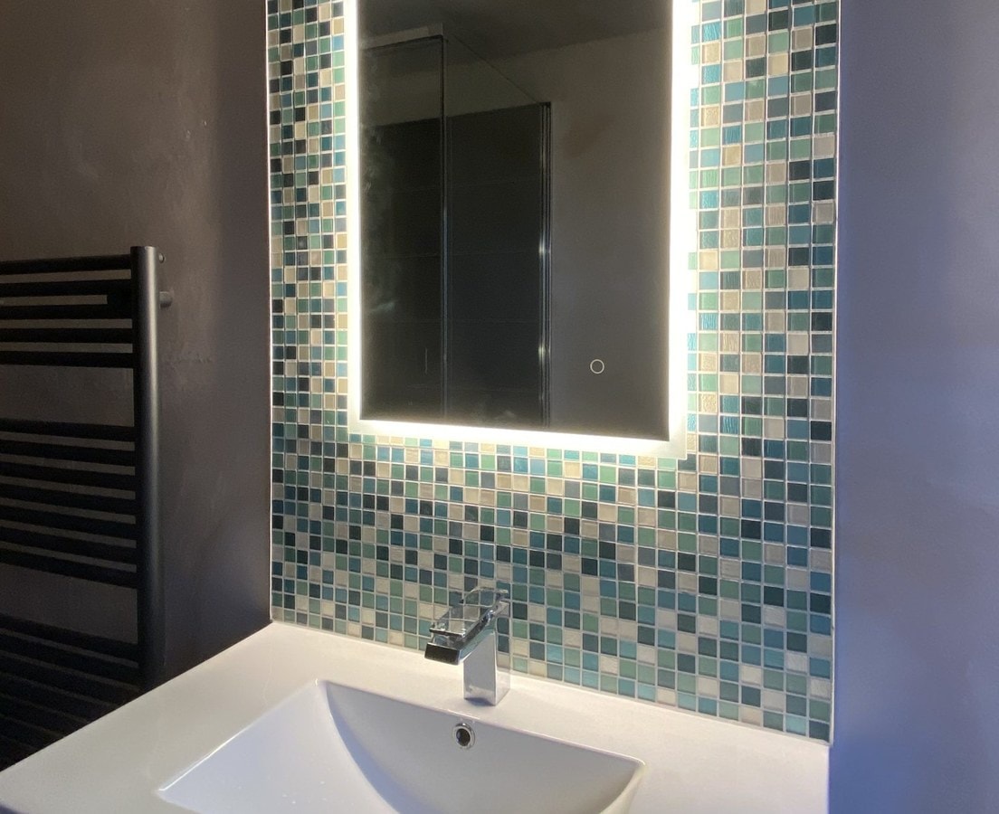 Lighted mirror over a vanity sink on a wall of blue-green glass mosaic tile
