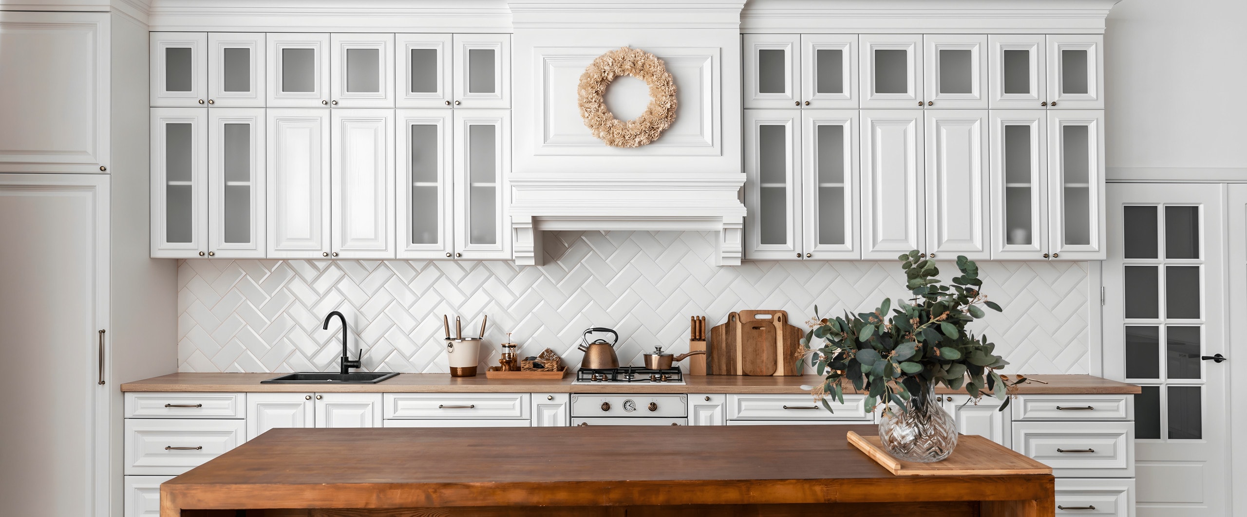 Wall of white raised-panel cabinets with glass-front uppers and a paneled range hood, behind a solid wood island
