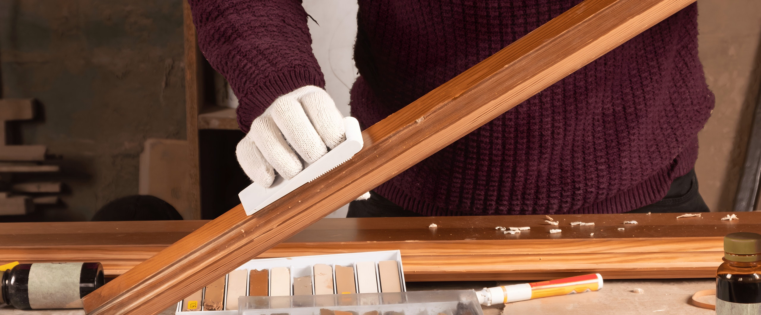 Carpenter in work gloves filling and smoothing a length of stained wood molding at the bench