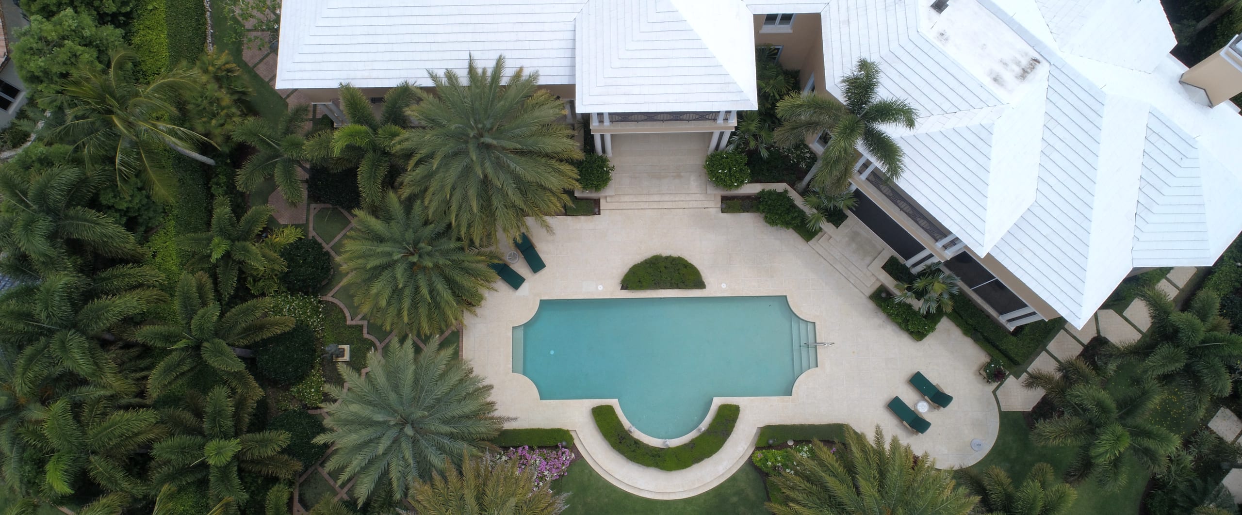 Aerial view of a courtyard pool with a stone deck, lounge chairs, and palm trees beside a white-roofed Florida home