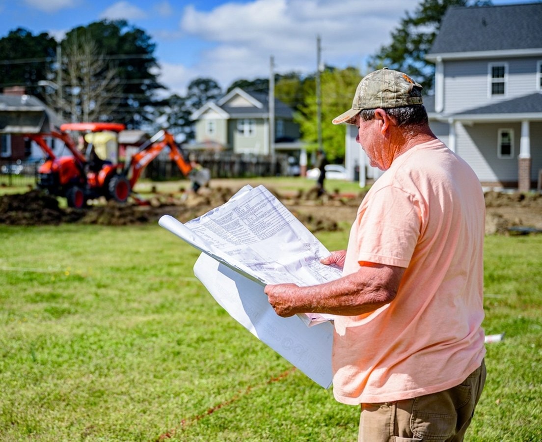 Builder reading site plans on a staked-out lot while a backhoe digs behind him