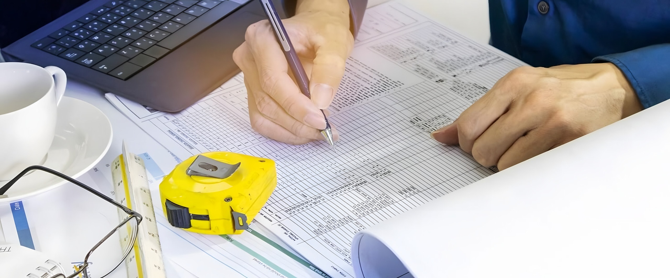 Hands filling in a printed project worksheet beside a tape measure, scale ruler, and laptop