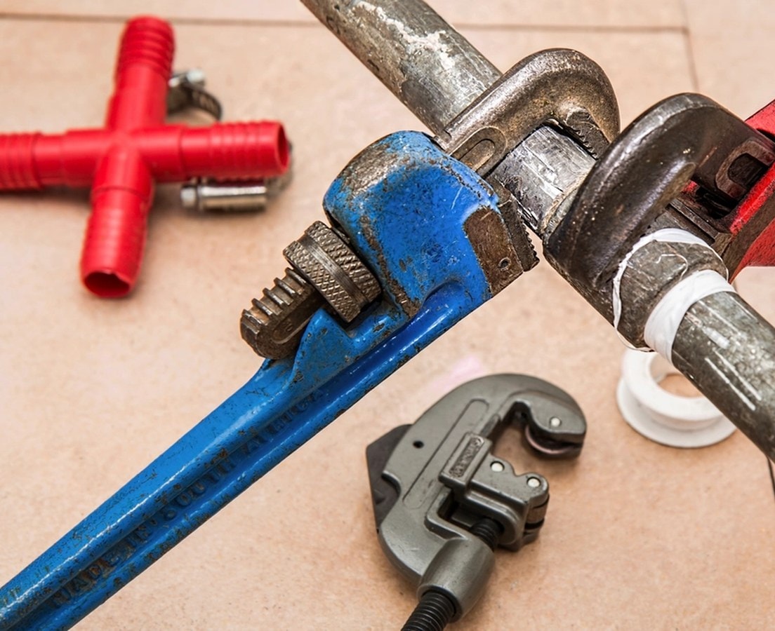 Pipe wrenches gripping a threaded supply pipe sealed with thread tape, beside a tubing cutter