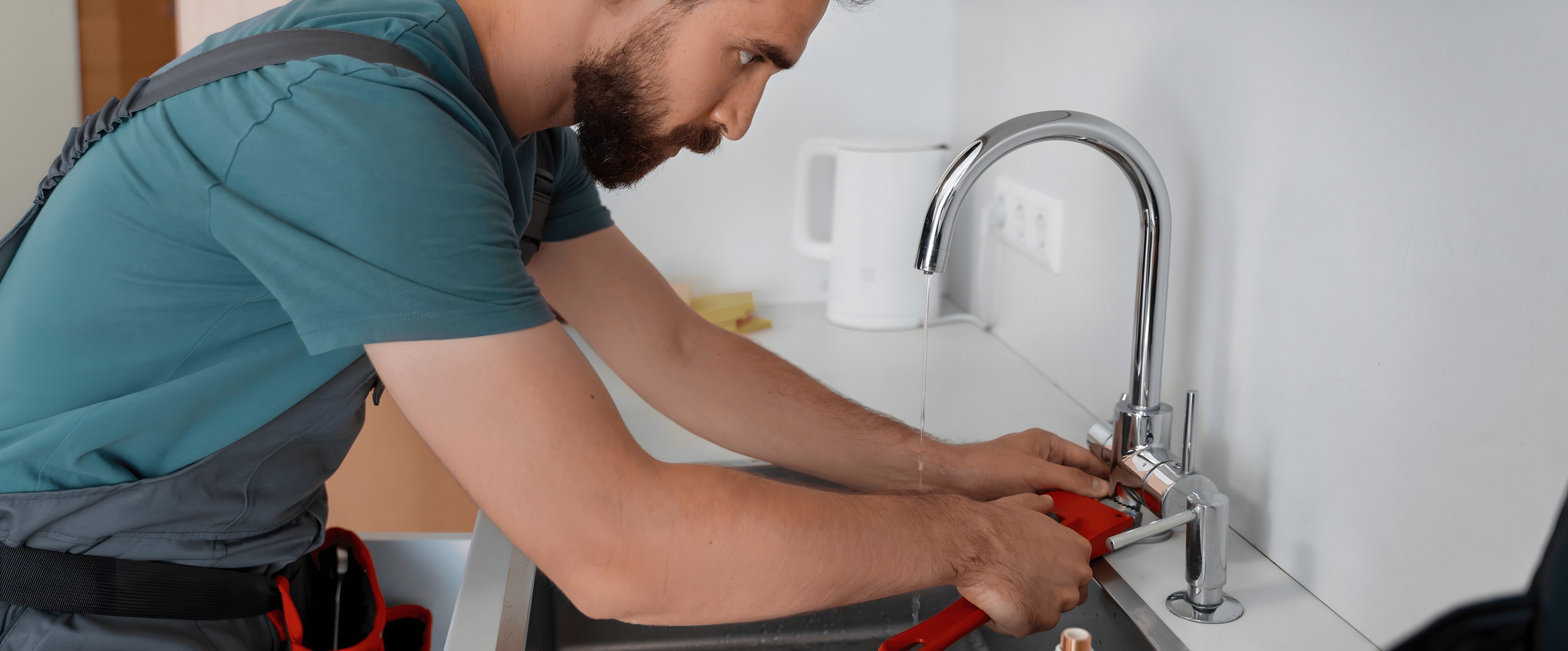 Plumber tightening a chrome kitchen faucet at the sink with a red pipe wrench