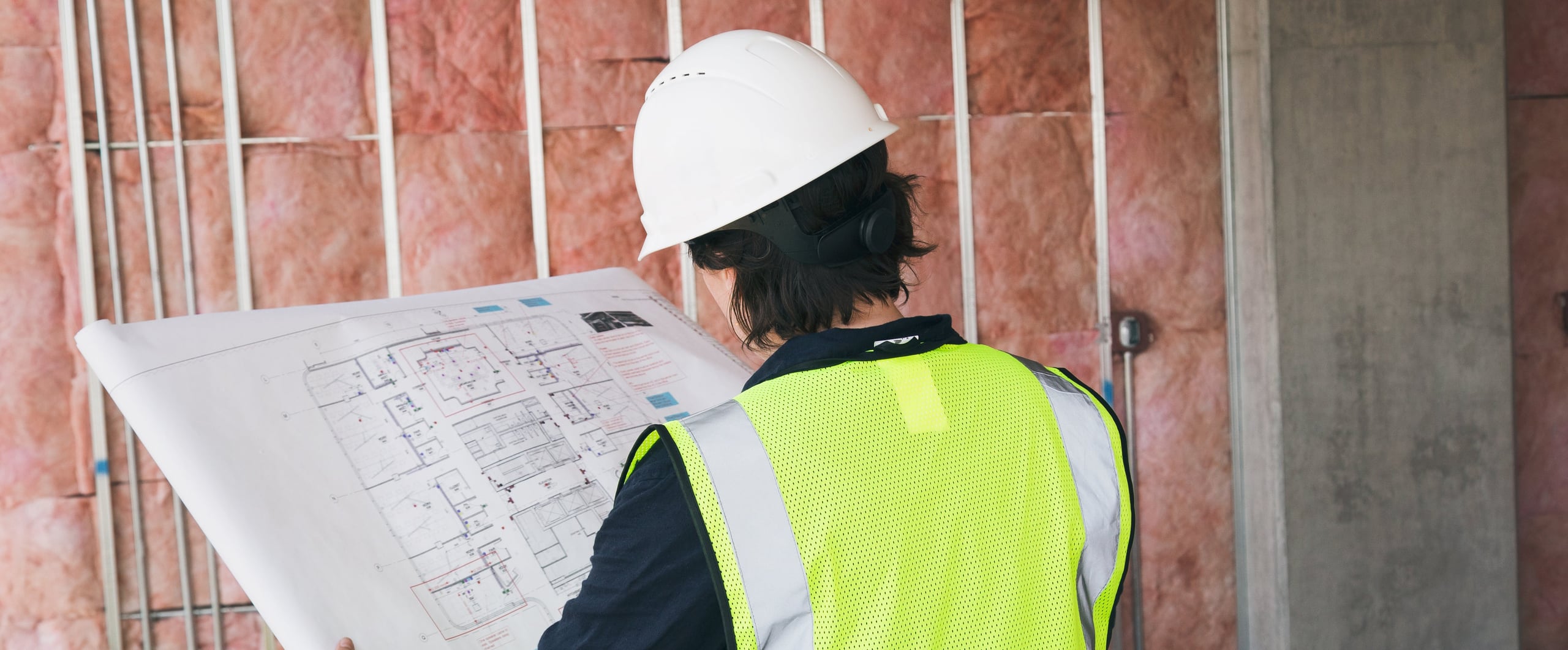 Project manager in a hard hat and safety vest checking floor plans against open stud walls and insulation before drywall