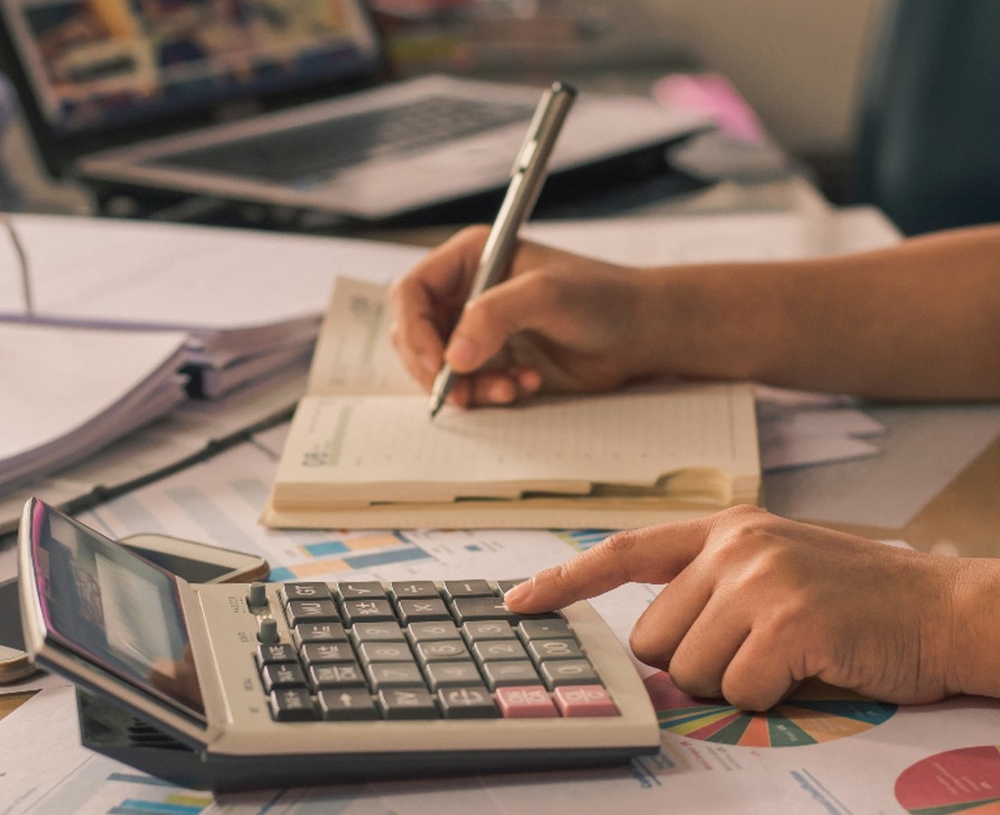 Hands working a desk calculator and writing in a planner beside binders and printed charts