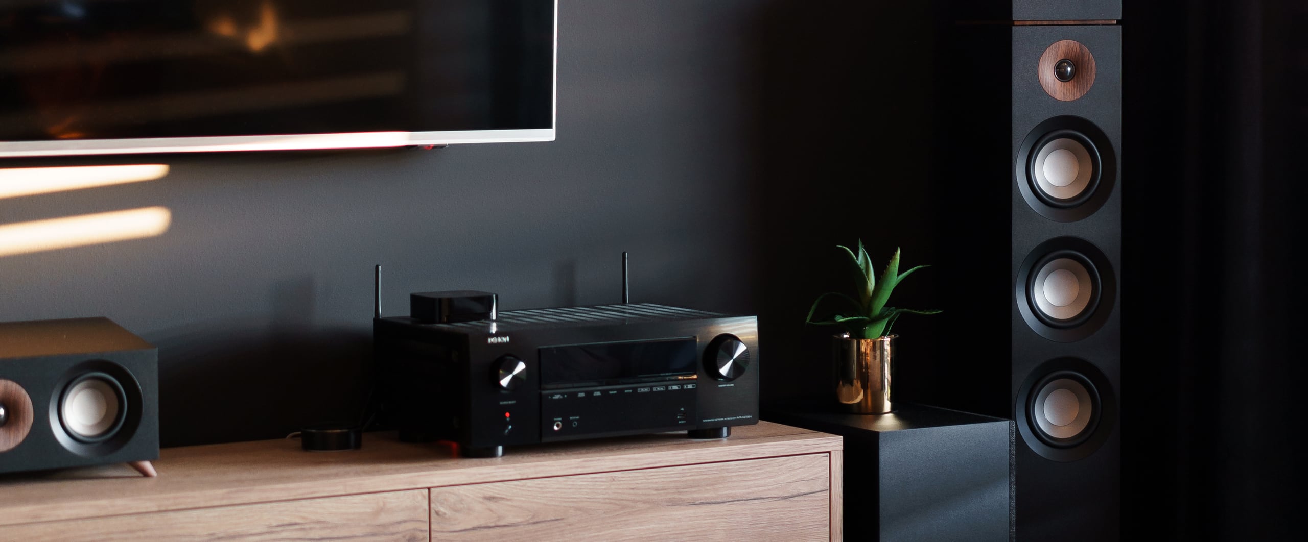 Media wall with a wall-mounted TV, an AV receiver, and center and tower speakers on a wood console, with no visible cables